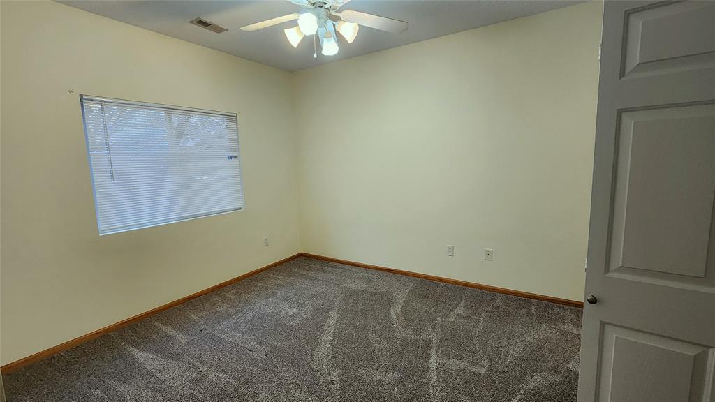 An empty room with beige walls and gray carpet. A window with blinds is on the left wall, and a ceiling fan with lights is in the center. A partially open door is on the right.