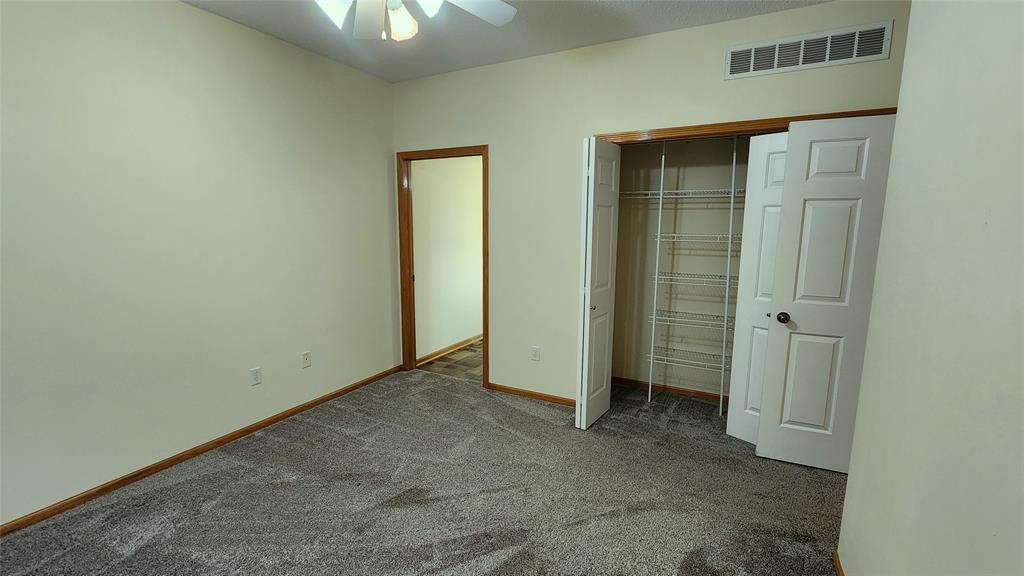 An empty room with beige walls and gray carpet. There's a ceiling fan with lights. The room has an open closet with white wire shelving and a wooden door leading to a hallway. Trim around the door and closet is wooden.