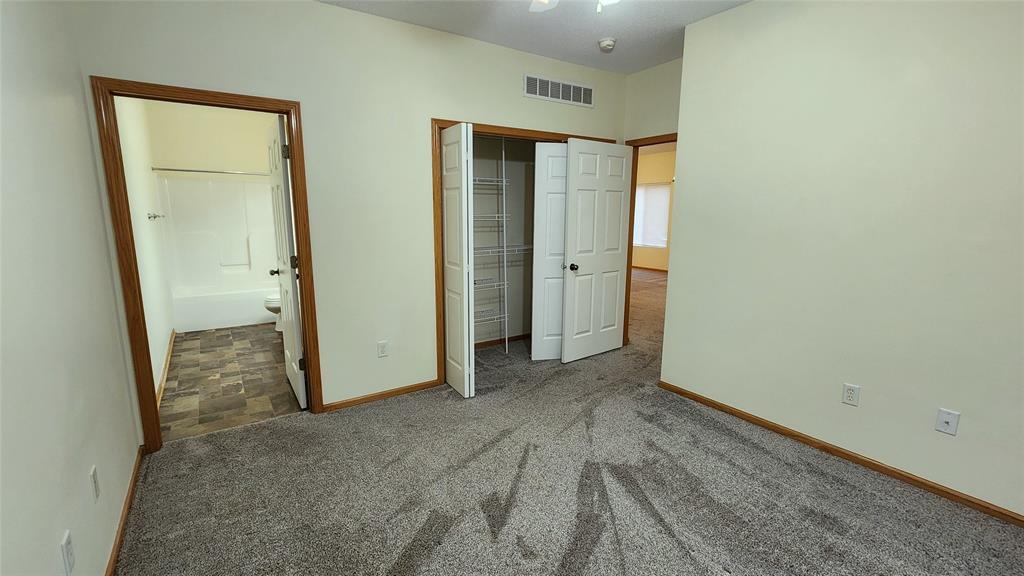 Empty room with light beige walls and carpeted floor. It features a closet with open white double doors, a door leading to a bathroom with a tub, and an open doorway to another carpeted room. Wood trim accents doors and baseboards.