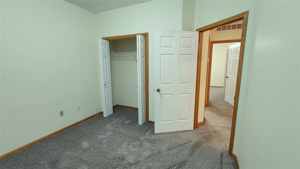 An empty room with light-colored walls and gray carpet. There is a closet with a white bifold door on the left and an open white door leading to a hallway with additional doors. The room has a simple ceiling and wood trim.