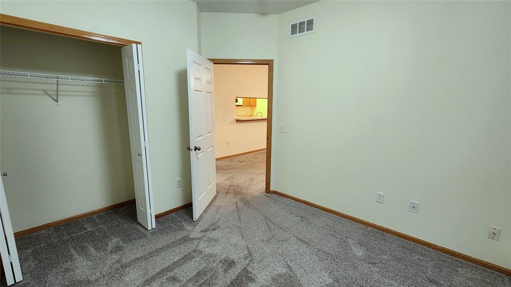 Empty room with light green walls, open closet on the left, and white door leading to a carpeted hallway. A view through the doorway shows a kitchen area with light wood cabinets and a countertop.