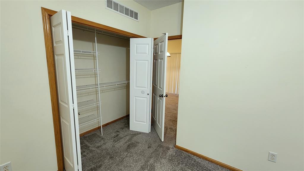 Empty room with light beige walls and gray carpet. White double-doors open to a closet with wire shelving on the left, and another door leading to a hallway on the right. Brown trim highlights door frames and baseboards.