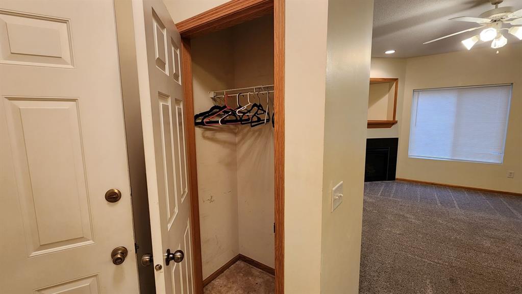 Open closet with empty hangers in an entryway, adjacent to a room with a ceiling fan and large window with blinds. The walls are cream-colored with wood trim, and the floor is carpeted. A portion of a fireplace is visible in the background.