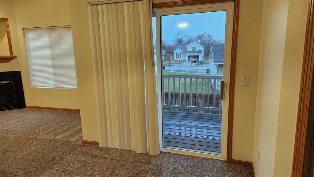 A room with carpeted flooring, a sliding glass door, and vertical blinds open to reveal a small wooden deck. Beyond the deck, a suburban street is visible with houses and trees in the background. A window with blinds is on the left wall.