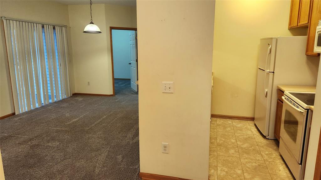 Interior view of an unfurnished apartment with a carpeted living area to the left, featuring vertical blinds on sliding doors. On the right is a tiled kitchen with a refrigerator, stove, microwave, and wooden cabinets. Walls are painted light yellow.