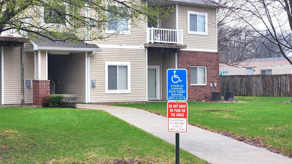 A pathway leads to an apartment building entrance. A blue disabled parking sign and a red sign below it, warning not to park on the grass, are posted at the center of the grassy yard. Trees and a wooden fence are in the background.