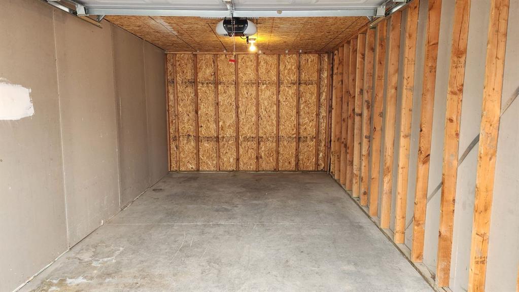 View of an empty garage with concrete floors and exposed wooden wall framing. The ceiling has an automatic door opener with a single light bulb turned on, illuminating the space. The walls are a mix of drywall and exposed wood.