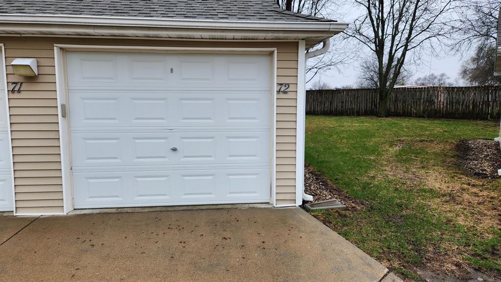 A closed white garage door with the number 72 is shown. The garage is attached to a house with beige siding. A wet concrete driveway leads to the door. A grassy yard and wooden fence are visible in the background. It's a cloudy day.
