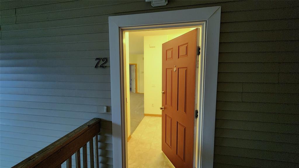 A slightly open brown door with the number 72 reveals a view into an empty room with yellowish lighting. The entrance is part of a wooden paneled exterior. A wooden railing is visible to the left of the door.