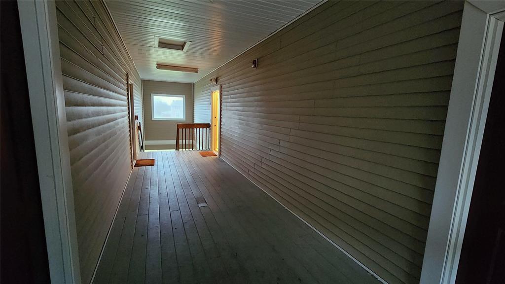 Dimly lit hallway with wooden floors and paneled walls. A softly glowing doorway is visible on the right, while natural light enters through a window ahead, creating a mix of warm and cool light tones.