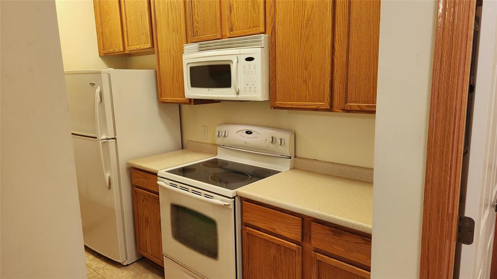 A small kitchen featuring wooden cabinets, a white refrigerator, a white stove with an oven, and a white microwave above it. The countertops are beige, and the floor has a light tile pattern.