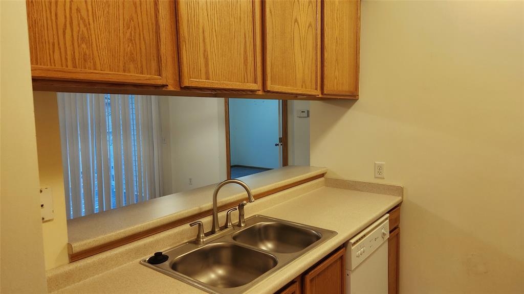 A kitchen features wooden cabinets above a light-colored countertop with a double stainless steel sink. A dishwasher is installed below the counter. A pass-through window opens to another room with vertical blinds.