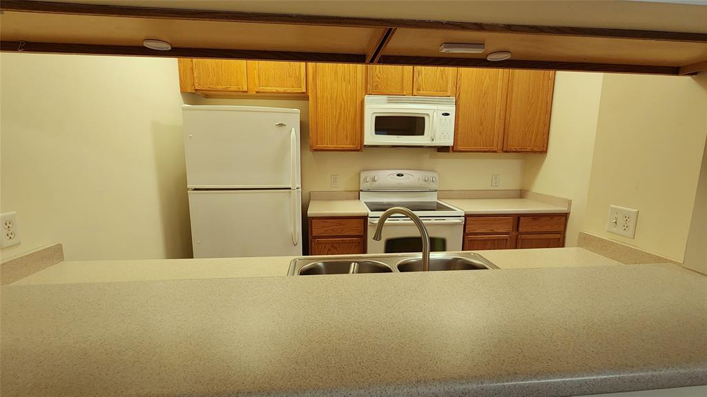 A kitchen with light wooden cabinets, white appliances including a refrigerator, microwave, and oven. A beige countertop with a double sink is in the foreground, viewed from an open pass-through window.