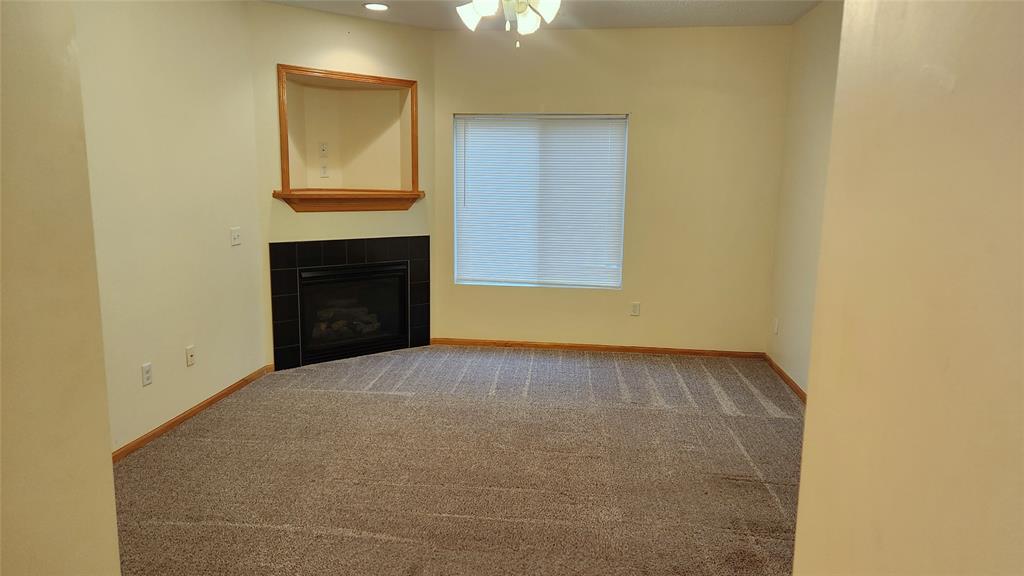 An empty living room with beige walls and carpeted floor. A large window with closed blinds is on the right wall. A corner fireplace with a wooden mantel and an inset shelf is in the corner. Ceiling fan above.