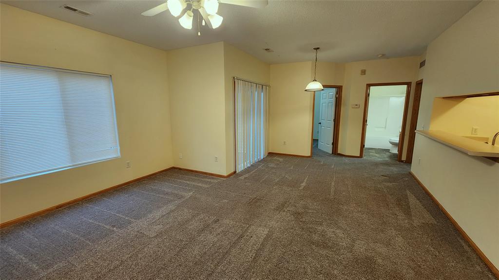 Spacious empty room with beige walls and carpeted floor. A ceiling fan hangs above, and there's a window with closed blinds on the left. In the back, a hanging light fixture, sliding glass doors, and an open door lead to another room.