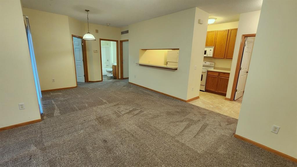 Spacious room with beige walls and brown carpet, featuring a central light fixture. A view into the kitchen shows wood cabinets and appliances. A small hallway leads to a visible open door with a glimpse of a bathroom through another door.