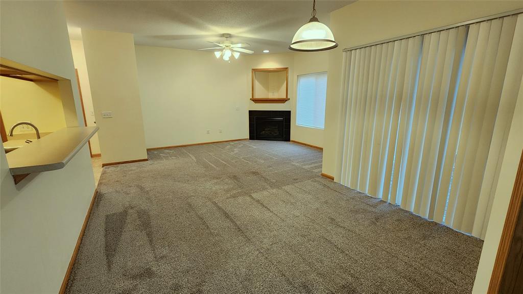 A vacant living room with beige walls and carpet. There's a ceiling fan with lights, a corner fireplace, a bright window, vertical blinds on sliding glass doors, and a partial view into the adjacent kitchen with a breakfast bar.