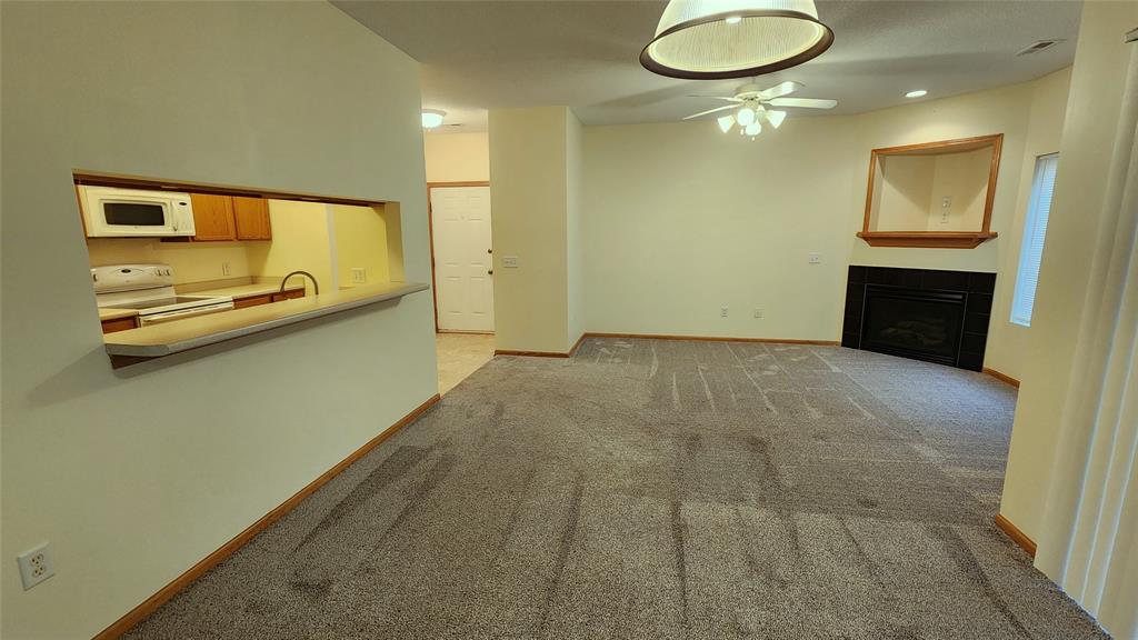 A spacious living room with gray carpet, a ceiling fan, and a corner fireplace. The room connects to an open kitchen via a serving hatch. The kitchen features wooden cabinets and white appliances. A round light fixture hangs from the ceiling.