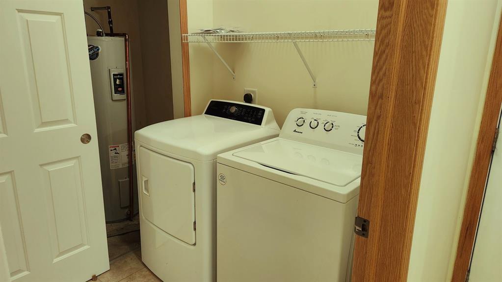A small laundry room with a white washing machine and dryer side by side. An electric water heater is visible in the corner. A wire shelf above holds some small items. The room has tan walls and light-colored tile flooring.