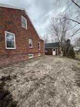 A brick house with several windows stands next to a bare yard with patchy dirt on Roche Street. Leafless trees surround the area, and a small detached building with a white door is visible in the background under a cloudy sky.