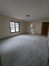 Empty carpeted room with beige walls, a ceiling light fixture, natural light from a window, a wooden door, and an open doorway leading to a kitchen with appliances—ideal real estate at 701 S Roche Street, Knoxville IA.