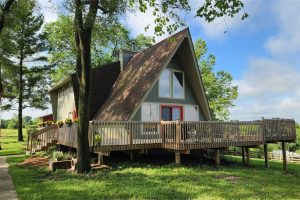 A-frame house with a large wooden deck, surrounded by green trees and grass under a blue sky with scattered clouds. The house has light gray siding, red front doors, and is elevated on stilts.