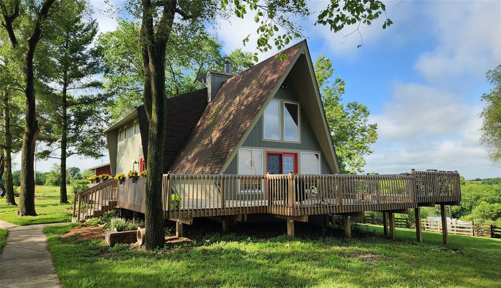 14434 US-65 Acreage 01 A-frame house with a large wooden deck, surrounded by green trees and grass under a blue sky with scattered clouds. The house has light gray siding, red front doors, and is elevated on stilts.
