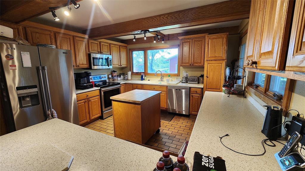 14434 US-65 Acreage 02 Spacious kitchen with wooden cabinets, stainless steel appliances, a central island, and large windows letting in natural light. Counters hold various small appliances and a tray of soda bottles.
