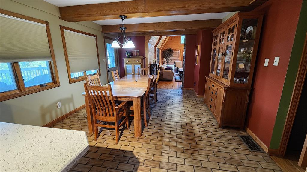 14434 US-65 Acreage 04 Dining room with wooden table and chairs, tile floor, china cabinet, and large windows. The room opens to a cozy living area with high ceilings and wood beams, decorated in warm earth tones.
