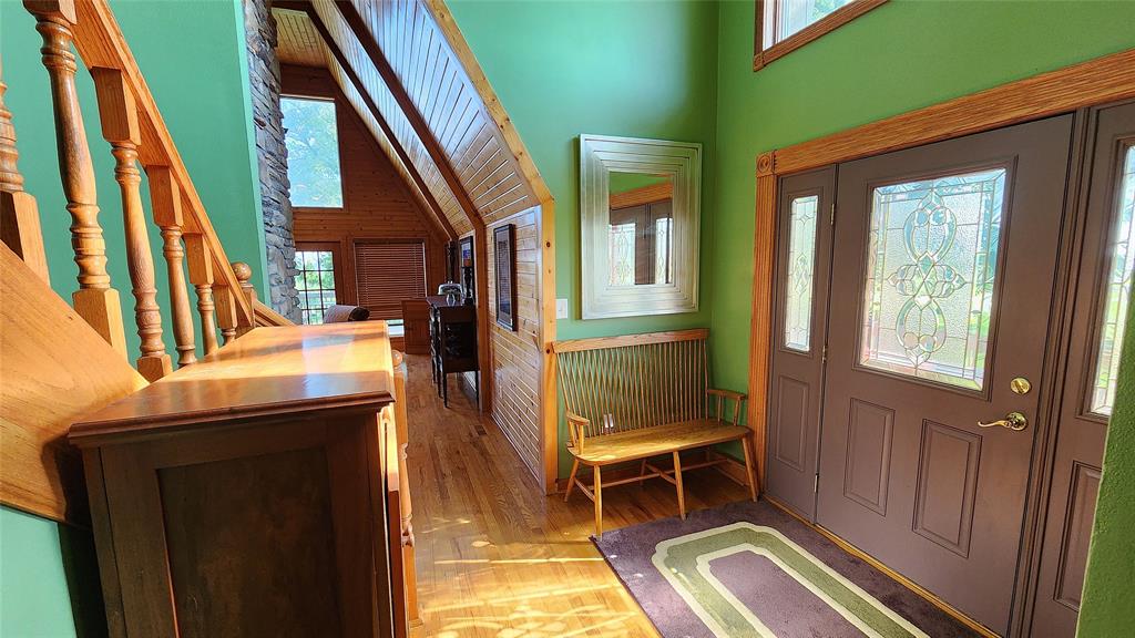14434 US-65 Acreage 05 Bright entryway with green walls, a large mirror, stained glass front door, wooden bench, staircase with wooden railing, hardwood floors, and a view into a warm, wood-paneled living area. Sunlight streams through the windows.