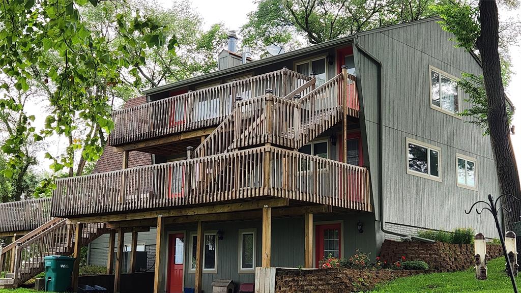 14434 US-65 Acreage 06 A three-story gray house with multiple red doors, large wooden decks on each level, and exterior staircases. The house is surrounded by trees and greenery, with a stone retaining wall and a green recycling bin visible.