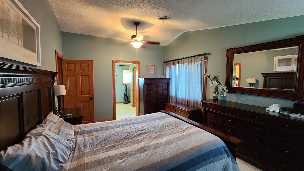 14434 US-65 Acreage 08 A cozy bedroom with a large bed, dark wood furniture, a mirror, and a window with sheer curtains letting in soft daylight. The walls are painted green, and a ceiling fan with a light hangs above.