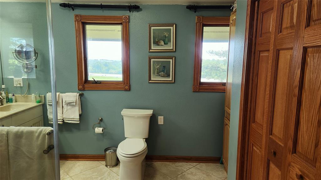 14434 US-65 Acreage 09 A bathroom with teal walls, two windows above a white toilet, two framed pictures between the windows, a vanity with a sink and towels on the left, and wood-paneled closet doors on the right.