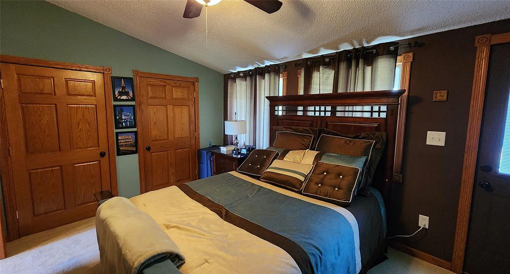 14434 US-65 Acreage 11 A cozy bedroom with a queen-sized bed featuring brown, blue, and beige bedding. The room has wood trim, two wooden doors, framed pictures on a green wall, a small dresser with a lamp, and tall curtained windows behind the bed.