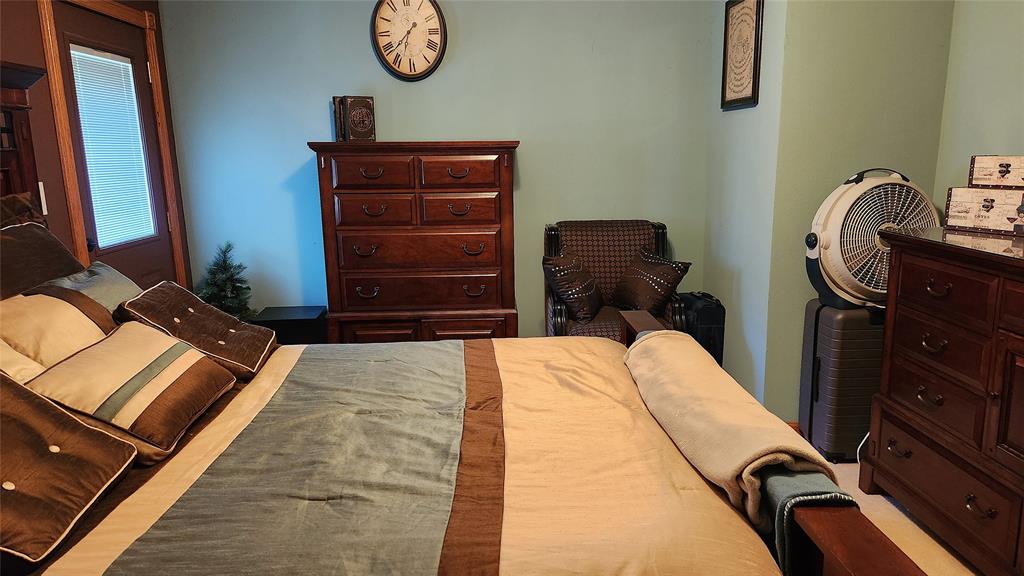 14434 US-65 Acreage 12 A neatly made bed with brown and tan bedding, a wooden dresser with a clock above it, a cushioned armchair, a large fan, and a small artificial tree in a cozy bedroom.