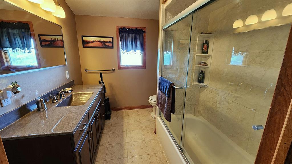 14434 US-65 Acreage 13 A bathroom with a double-sink vanity, granite countertop, dark cabinets, and a large mirror. There is a glass-enclosed shower-tub, toilet, and two small curtained windows with wall art above the towel rack.
