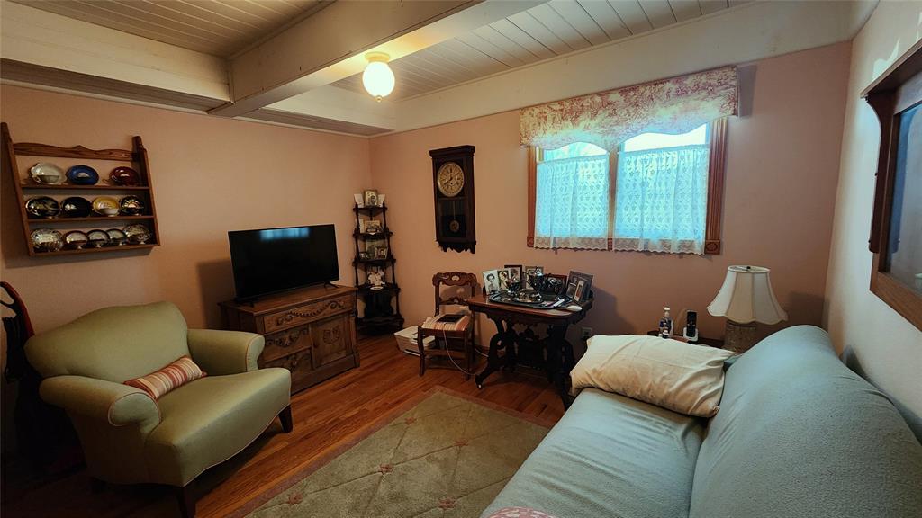 14434 US-65 Acreage 14 Cozy living room with pink walls, armchair, sofa, TV on a wooden cabinet, wall clock, shelves with teapots, a small table with photos, and a window with lace curtains letting in light.