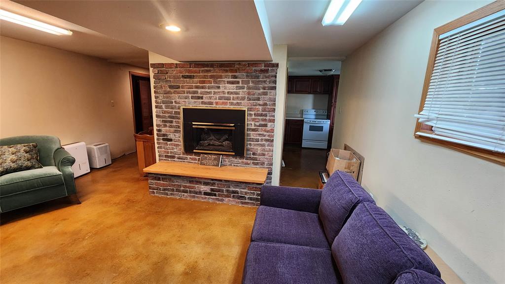 14434 US-65 Acreage 18 A cozy basement room with a brick fireplace, two covered couches, a wooden nightstand with a lamp, yellowish polished concrete floor, and a window with blinds letting in natural light.