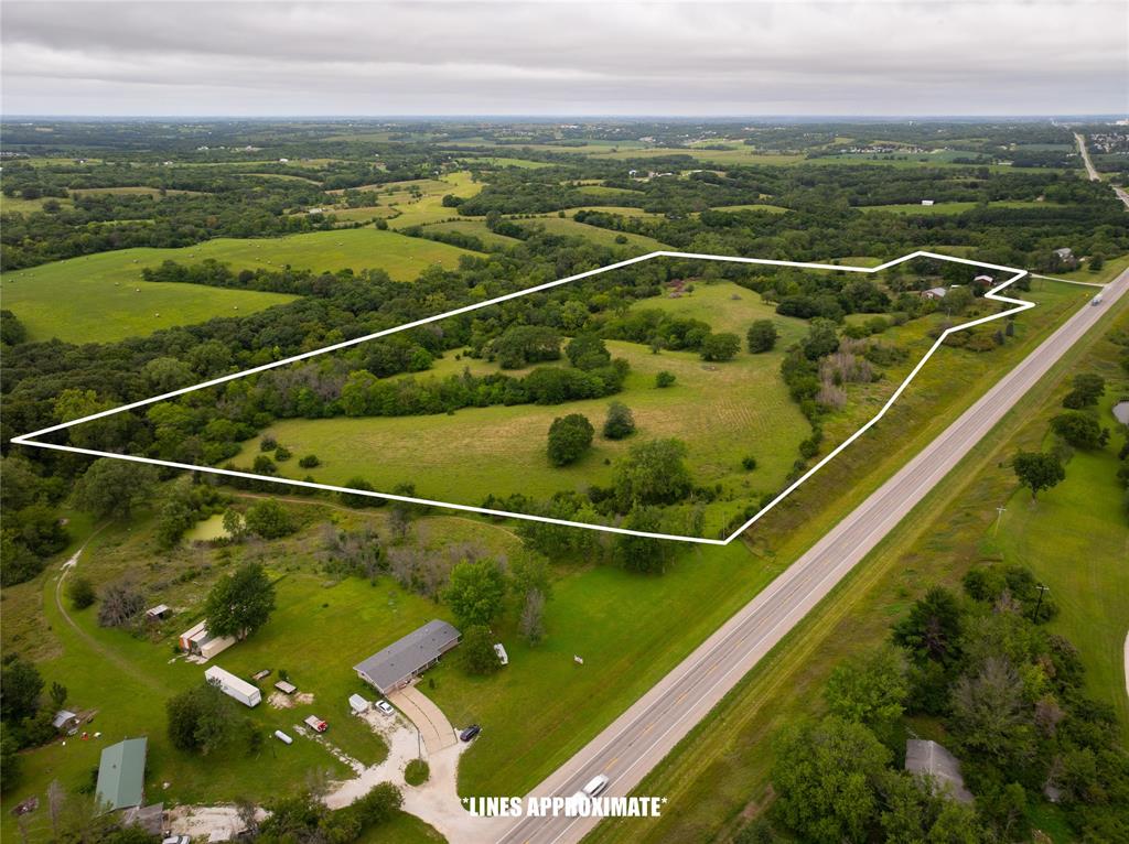 14434 US-65 Acreage 24 Aerial view of a large, open, grassy property outlined in white, bordered by trees and a road on one side, with scattered buildings and lush green landscape in the background.
