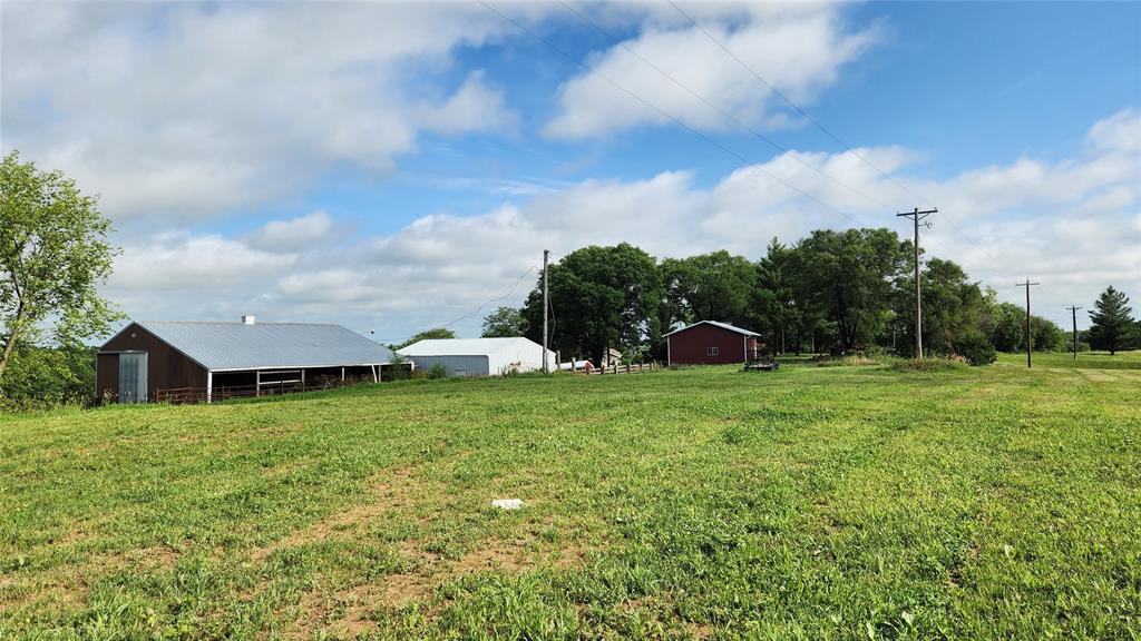 14434 US-65 Acreage 25 A grassy field with three red barns and utility poles, trees lining the background, and a partly cloudy blue sky overhead.