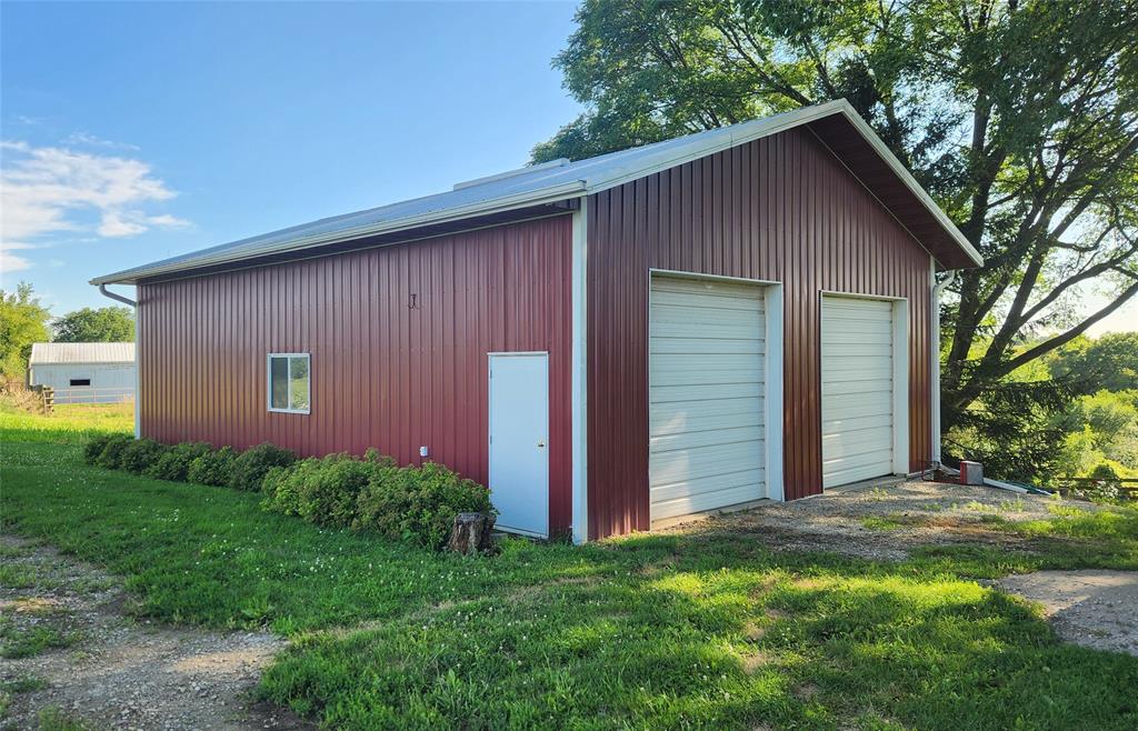 14434 US-65 Acreage 26 A red metal barn with two large white garage doors, a small entry door, and a window, surrounded by greenery and trees on a sunny day.