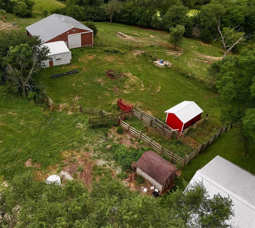default Aerial view of a rural farm with several fenced areas, a small red barn, a larger white-roofed barn, a wooden shed, green grass, and trees surrounding the property.