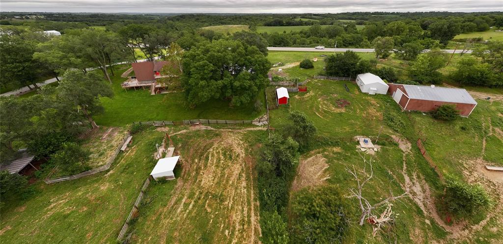 default Aerial view of a rural property with a house, green lawns, trees, fenced areas, red barns, sheds, and open fields under a cloudy sky. A road runs behind the property, with rolling hills in the distance.