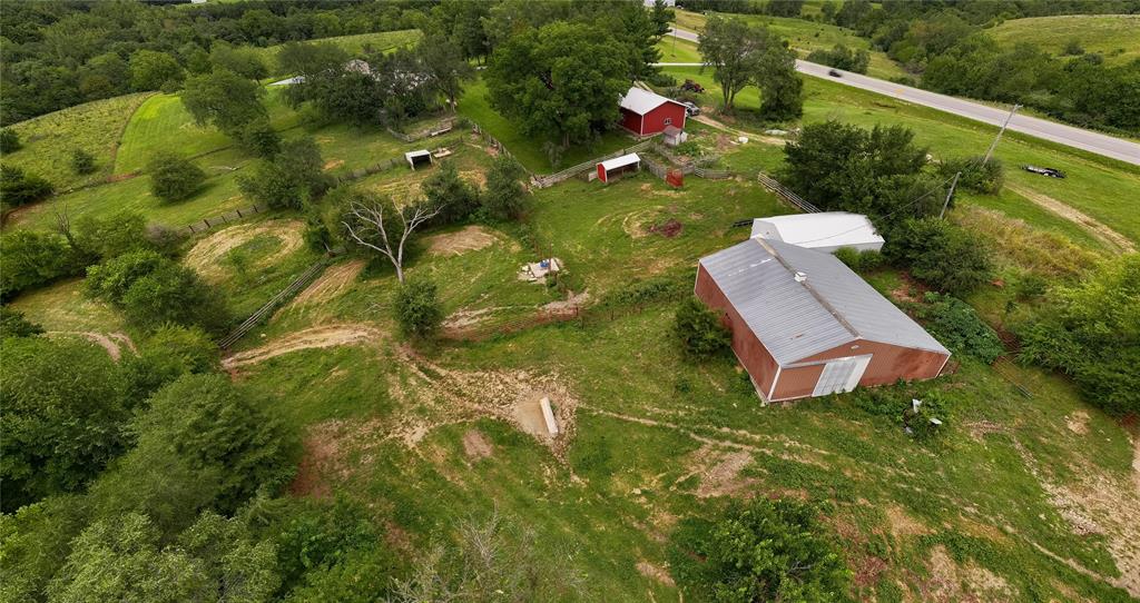 default Aerial view of a rural landscape featuring a large red barn, a smaller red building, scattered trees, fenced areas, and dirt paths surrounded by green fields near a road.