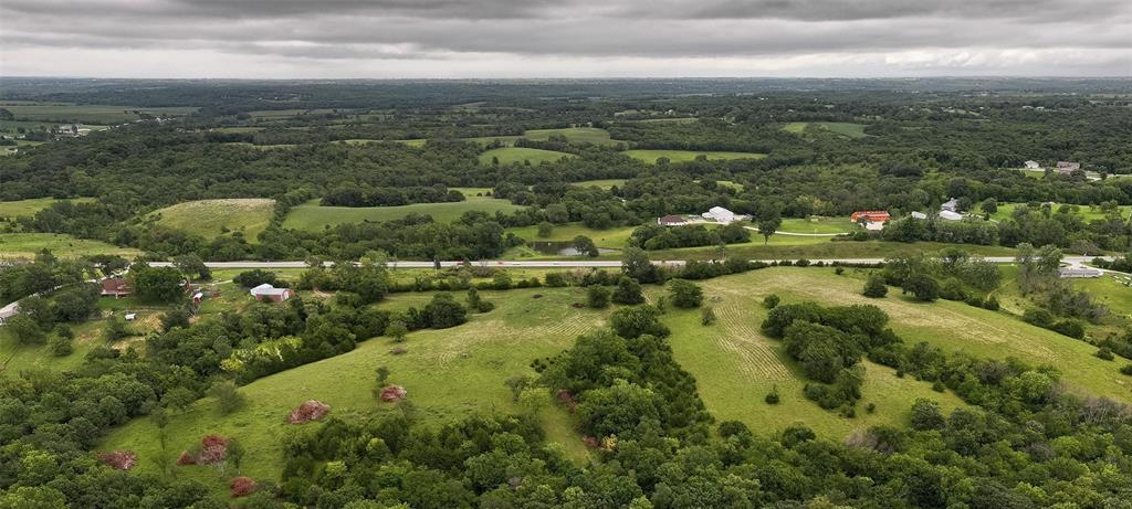 default Aerial view of a rural landscape with rolling green hills, scattered trees, houses, fields, and a road under a cloudy sky.