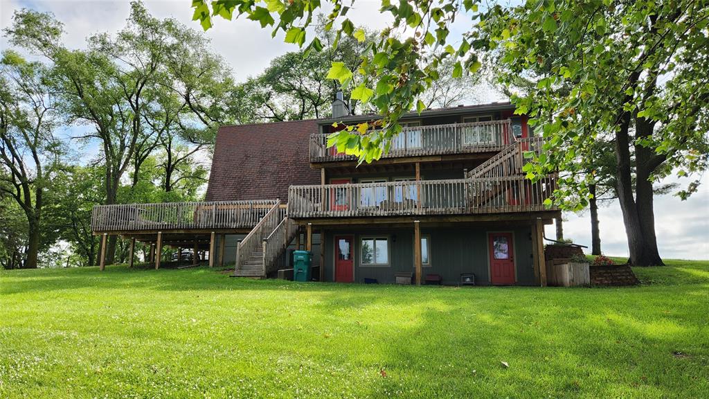 14434 US-65 Acreage 31 A large, wooden, three-story house with wraparound decks and stairs stands on a grassy lawn, surrounded by tall leafy trees under a partly cloudy sky.