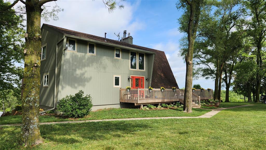 14434 US-65 Acreage 32 A modern two-story house with sage green siding, a dark sloped roof, and a bright red front door, surrounded by grass, trees, and landscaping, under a blue sky with scattered clouds.
