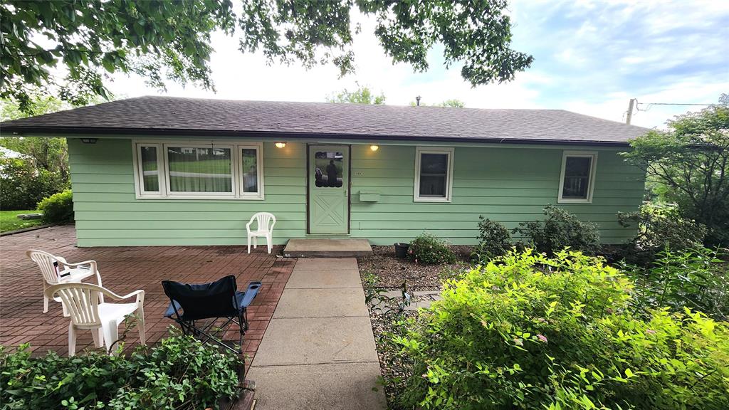 202 E Main St 01 A small, light green house with a dark roof and white trim, featuring a front door and windows. Patio chairs and a paved walkway lead to the entrance, surrounded by bushes and greenery.
