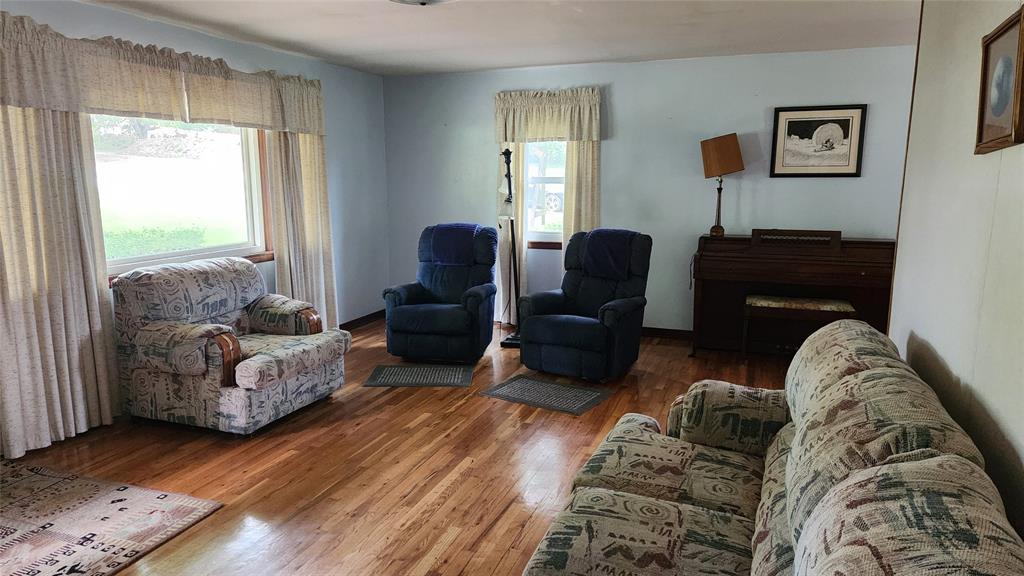202 E Main St 02 A living room with wood floors, patterned sofa and armchair, two blue recliners, large windows with sheer curtains, a piano in the corner, and framed art on light blue walls.