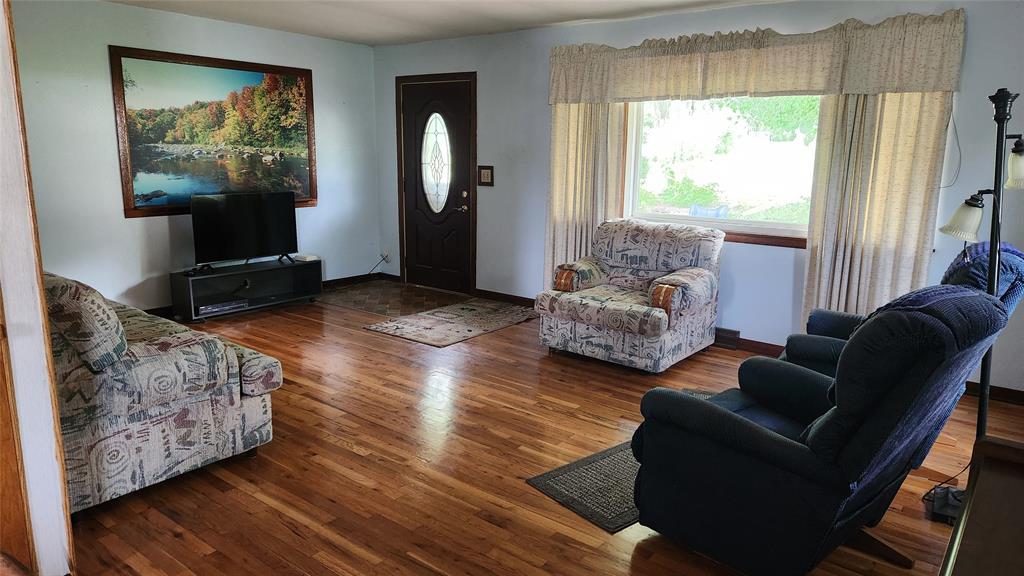 202 E Main St 03 A living room with hardwood floors, a patterned sofa, two armchairs, a TV on a stand, a floor lamp, a large window with curtains, and a door with an oval glass window. A landscape painting hangs above the TV.